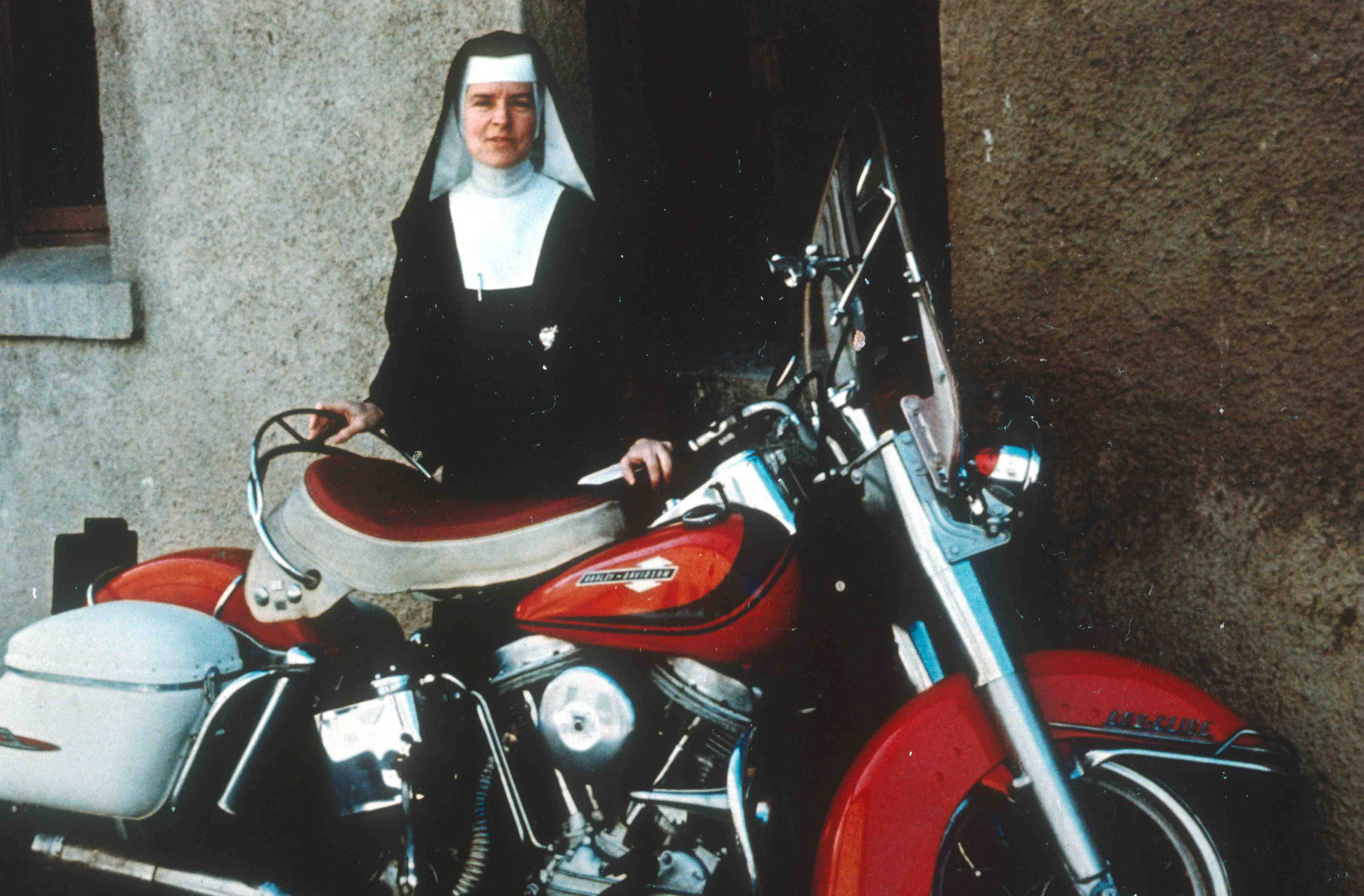 Sister Corita Kent, a nun, sitting behind a red motorbike